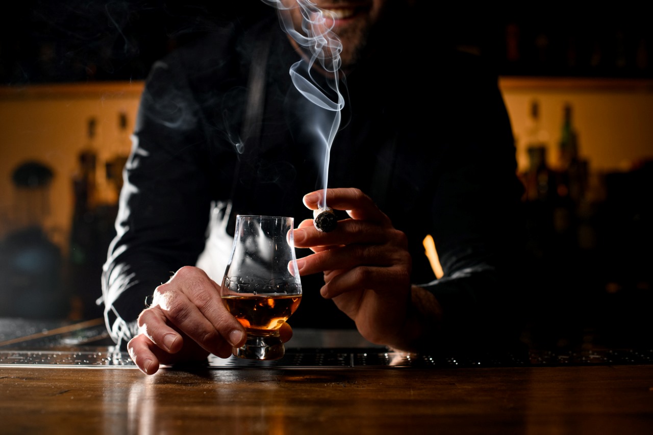 view of male hands holding a glass of strong alcoholic drink and smoking cuban cigar